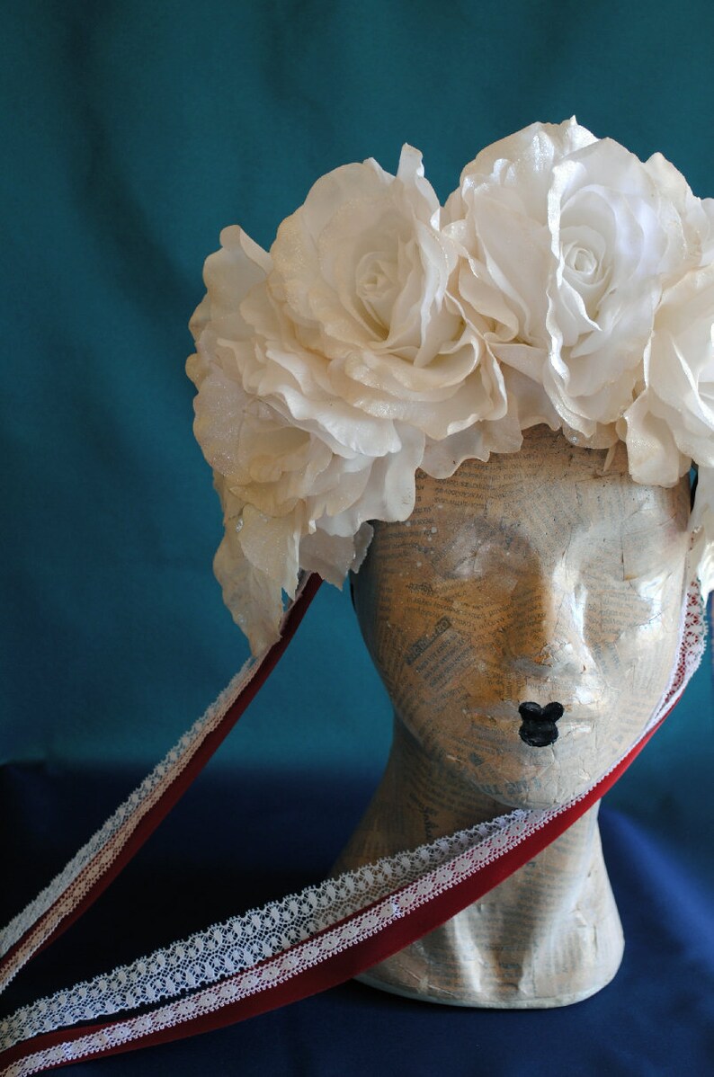 May include: A headpiece featuring a cluster of large, white fabric roses with a subtle shimmer. The floral arrangement is attached to a mannequin head covered in newsprint, with two long, dark red velvet ribbons trimmed in white lace hanging down.