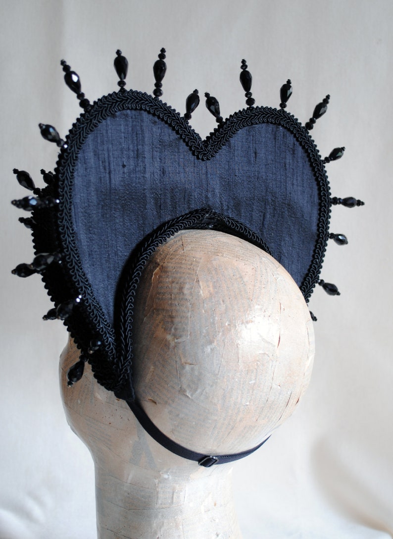 May include: A black fabric heart-shaped headpiece with black bead embellishments. The headpiece has a black elastic band for securing it to the head.