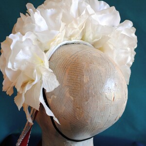 May include: A white floral headband featuring a dense arrangement of large, layered fabric rose blossoms attached to a thin, dark-colored band. A long, thin red ribbon with a white lace trim hangs down from the side of the floral headpiece.