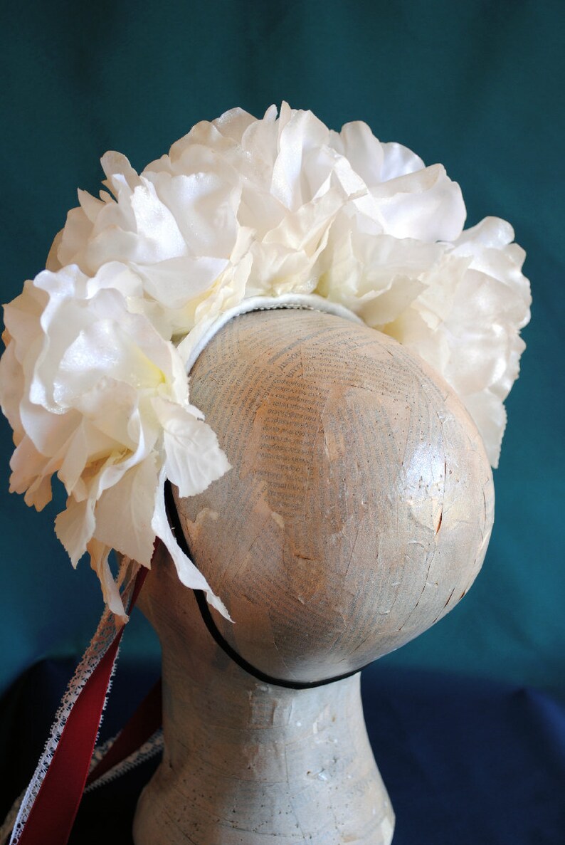 May include: A white floral headband featuring a dense arrangement of large, layered fabric rose blossoms attached to a thin, dark-colored band. A long, thin red ribbon with a white lace trim hangs down from the side of the floral headpiece.
