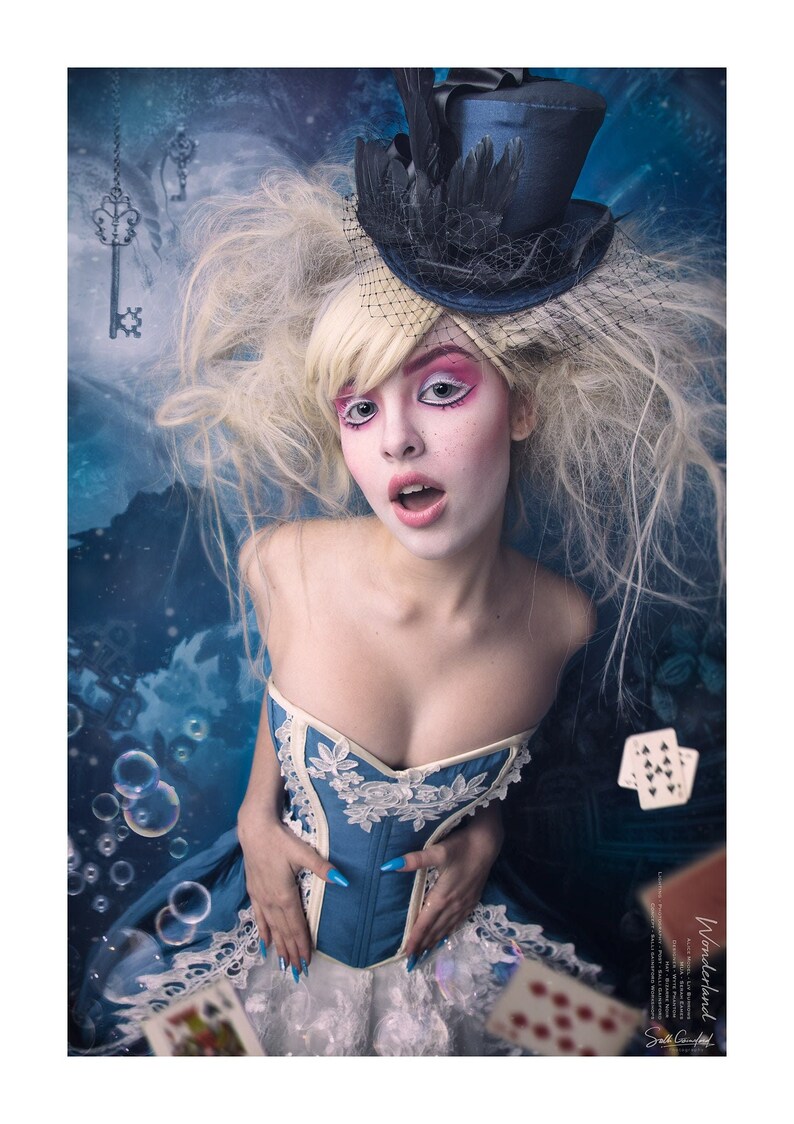 May include: A woman with blonde hair and blue eyes wears a blue and white corset with lace detailing. She is wearing a blue top hat with a black veil and has dramatic makeup. The background is a blue and white abstract design with playing cards and bubbles.