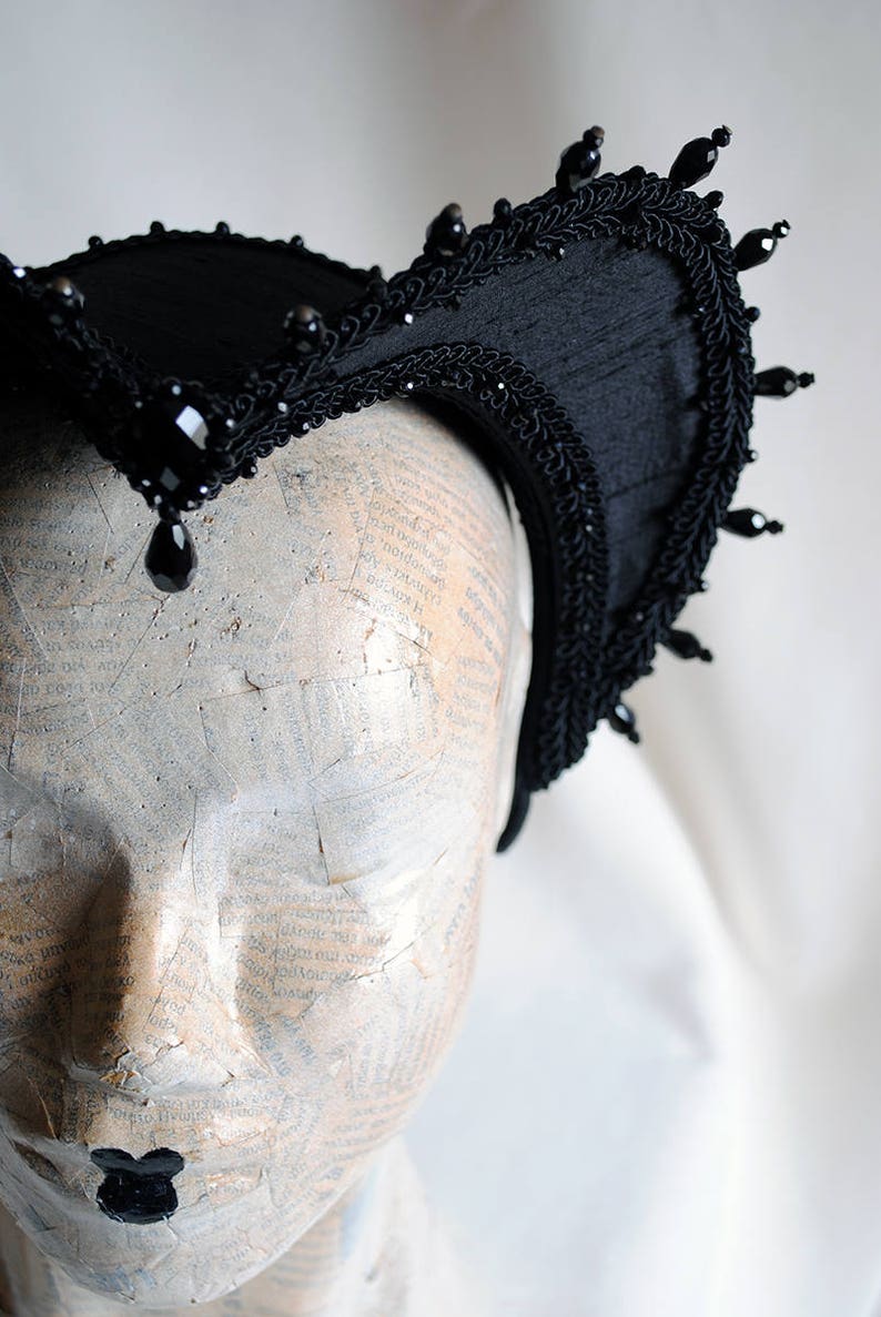 May include: A black headpiece featuring a structured, curved frame adorned with intricate braided trim and faceted black beads. A prominent, central teardrop-shaped black crystal hangs from the front, while smaller beads line the outer edge.