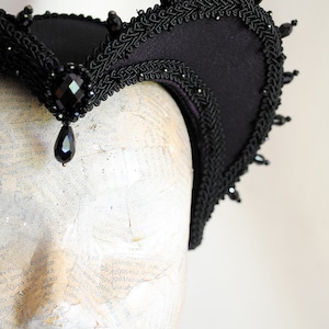 May include: A black fabric headband with a curved shape and a beaded trim. The headband is decorated with black beads and has a teardrop-shaped black jewel hanging from the center.
