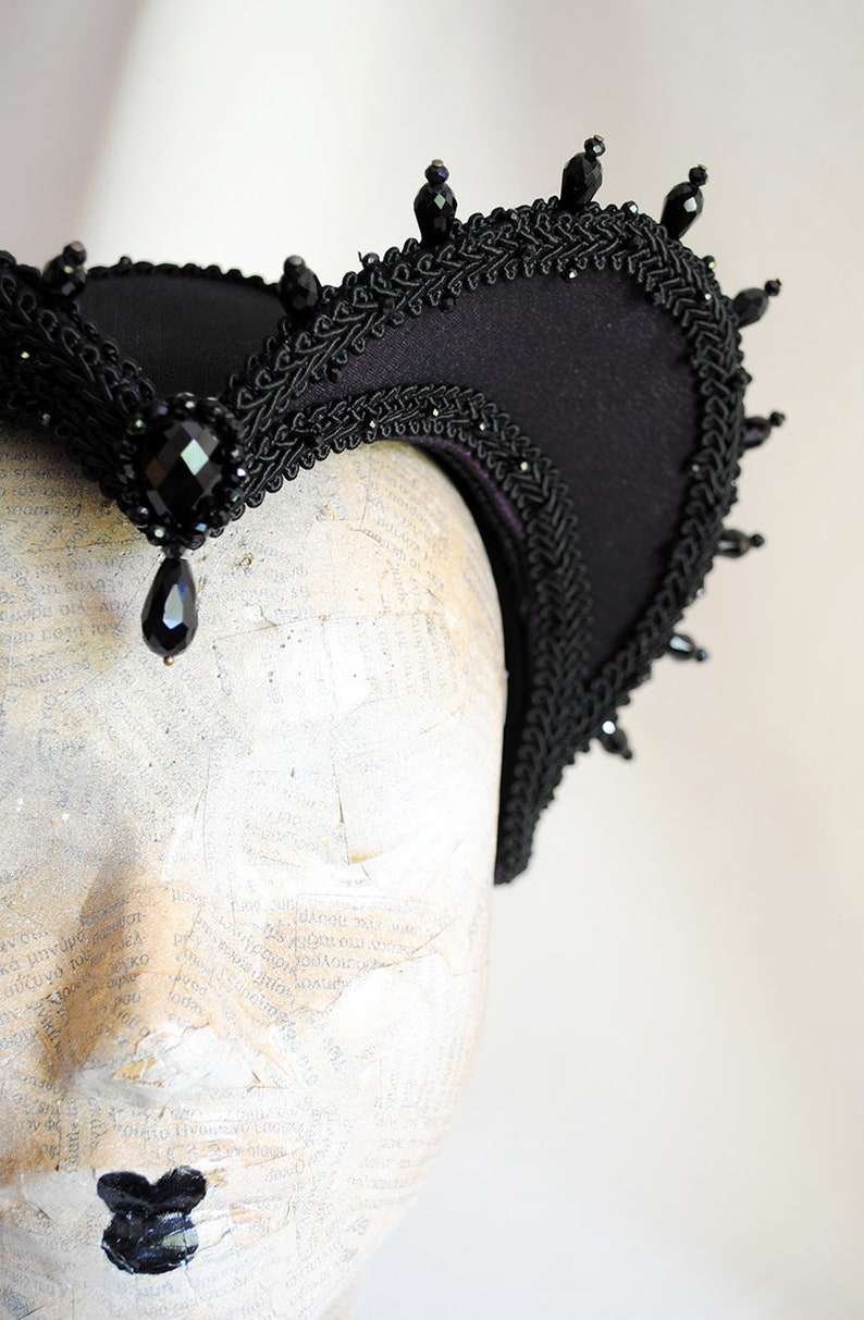 May include: A black fabric headband with a curved shape and a beaded trim. The headband is decorated with black beads and has a teardrop-shaped black jewel hanging from the center.