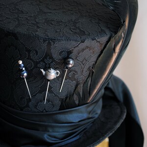 May include: A black top hat with a black feather and a black satin band. The hat is decorated with four pins, including a teapot, a bead, and two black buttons.
