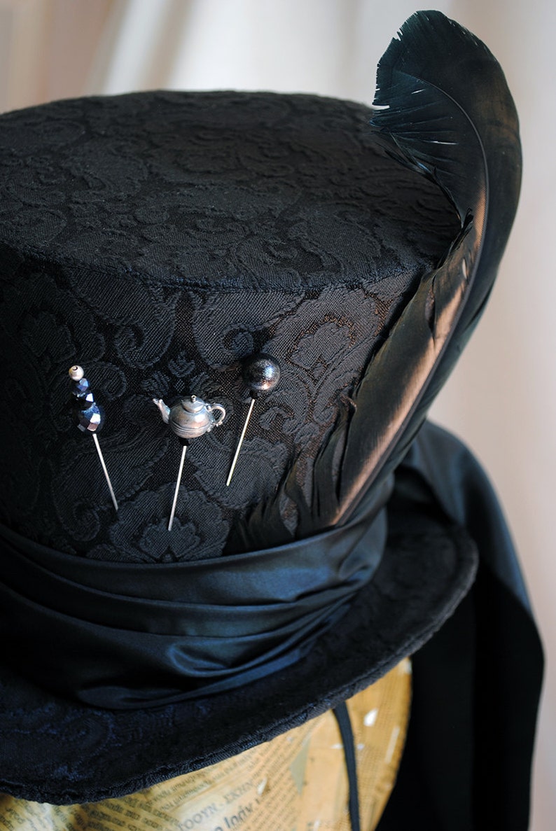 May include: A black top hat with a black feather and a black satin band. The hat is decorated with four pins, including a teapot, a bead, and two black buttons.