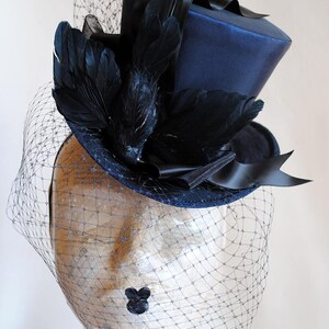 May include: A navy blue top hat with a black satin ribbon and black feathers. The hat is adorned with a black veil.