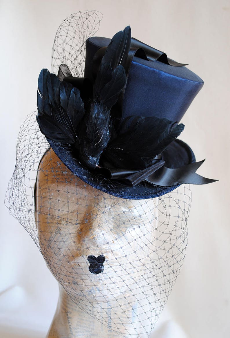 May include: A navy blue top hat with a black satin ribbon and black feathers. The hat is adorned with a black veil.