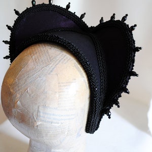May include: A black velvet hat with a scalloped brim and black bead trim. The hat is on a white mannequin head.