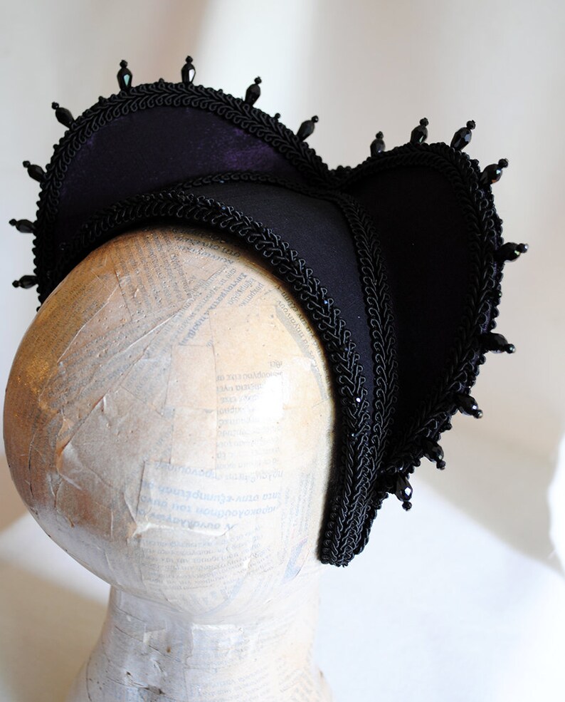 May include: A black velvet hat with a scalloped brim and black bead trim. The hat is on a white mannequin head.
