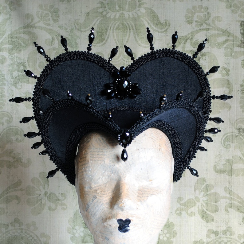 May include: A black heart-shaped headpiece with black bead embellishments. The headpiece is made of fabric and has a pointed crown.