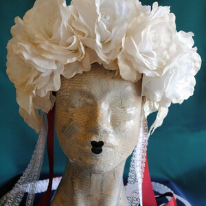 May include: A headpiece featuring a cluster of large, off-white fabric roses arranged in a crown shape. The headpiece includes long, dangling red ribbons and white lace trim accents, displayed on a mannequin head covered in vintage newspaper print.