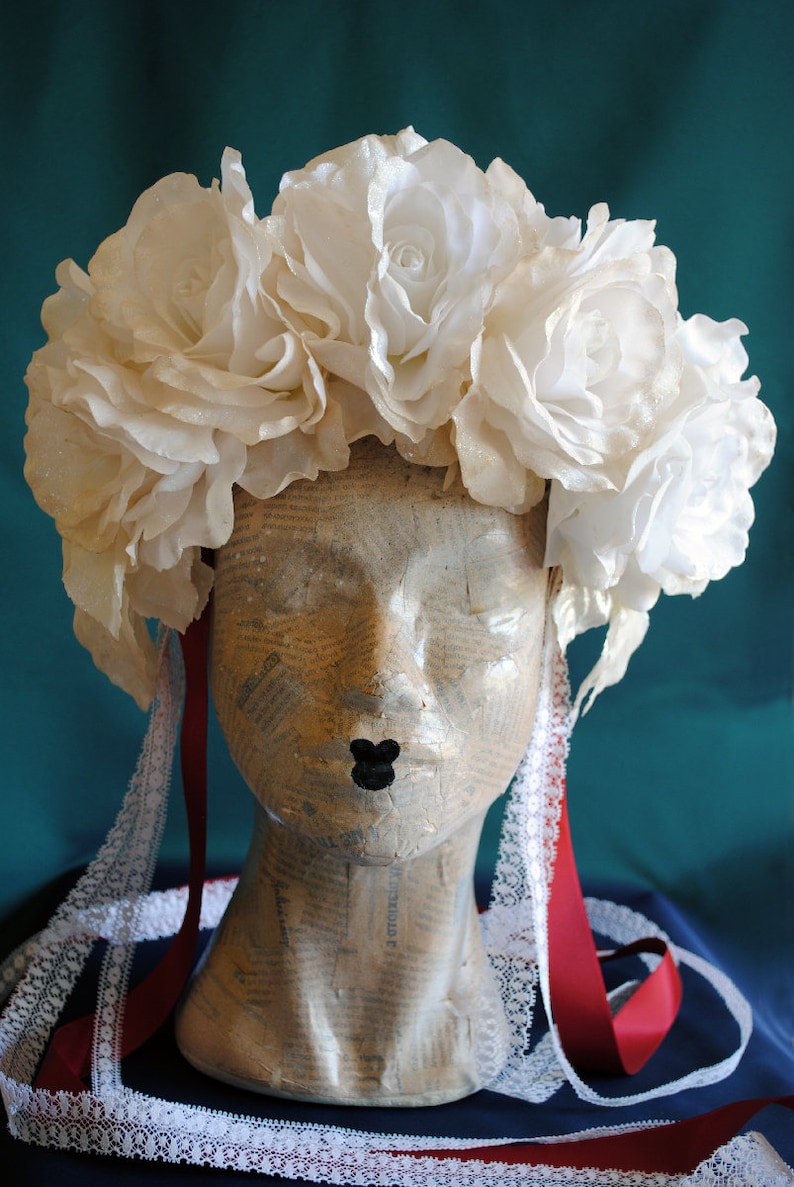 May include: A headpiece featuring a cluster of large, off-white fabric roses arranged in a crown shape. The headpiece includes long, dangling red ribbons and white lace trim accents, displayed on a mannequin head covered in vintage newspaper print.