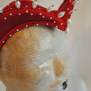 May include: A deep red headpiece featuring a structured, curved silhouette adorned with intricate red braided trim and rows of white teardrop-shaped faux pearls. The headpiece is displayed on a mannequin head covered in newsprint text.