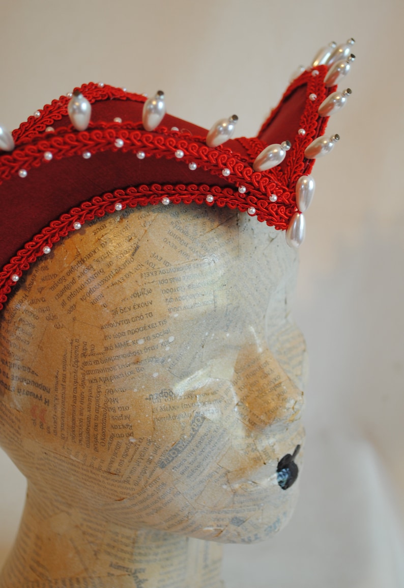 May include: A deep red headpiece featuring a structured, curved silhouette adorned with intricate red braided trim and rows of white teardrop-shaped faux pearls. The headpiece is displayed on a mannequin head covered in newsprint text.