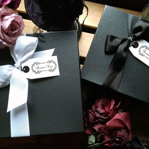 May include: Two square black gift boxes, one tied with a white ribbon and the other with a black ribbon. Each box features a white rectangular tag with the text Bizane Noir in a decorative script font, attached by a black string to the ribbon.