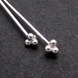 May include: A pair of silver-colored drop earrings. Each earring features a cluster of three small, round beads at the bottom of a thin, straight post. The earrings are displayed against a dark gray background.