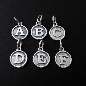 May include: Six silver-toned letter charms, A through F, each within a circular frame. The letters are raised against a textured background, with a small loop at the top for attachment. The charms are displayed against a black backdrop.