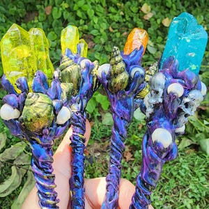 May include: Four handcrafted wands featuring iridescent purple and blue twisted handles. Each wand is topped with a unique crystal point in yellow, orange, or blue, accented with small white pearls, metallic gold or silver shells, and skulls.