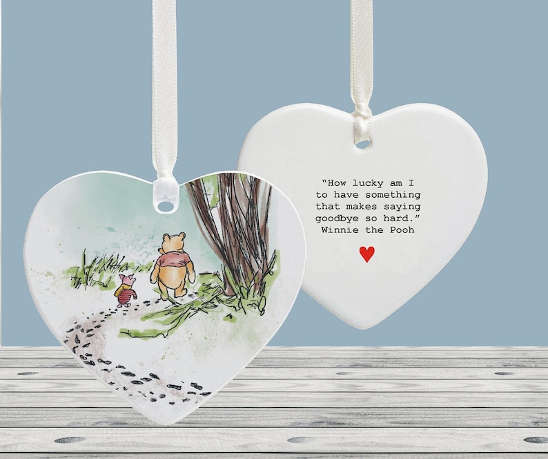 Peut inclure: Ornement en c&eacute;ramique blanche en forme de c&oelig;ur avec une illustration de Winnie l'ourson et Porcinet d'un c&ocirc;t&eacute; et une citation de Winnie l'ourson de l'autre. La citation est : "Quelle chance j'ai d'avoir quelque chose qui rend les adieux si difficiles." Winnie l'ourson.