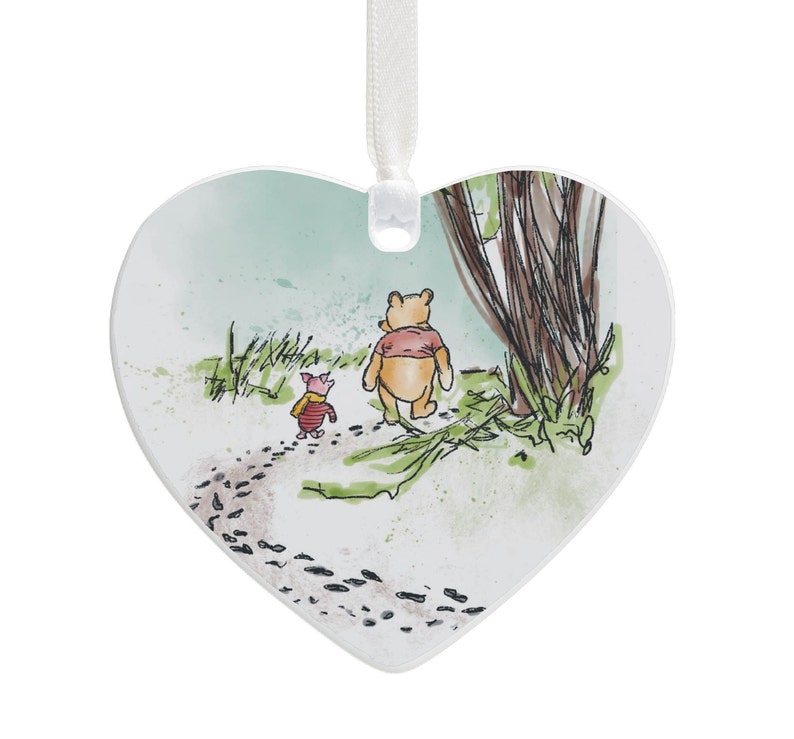 Peut inclure: Une d&eacute;coration en c&eacute;ramique blanche en forme de c&oelig;ur avec une illustration aquarelle de Winnie l'ourson et Porcinet marchant dans les bois. La d&eacute;coration a un ruban pour l'accrocher.