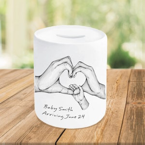 May include: White ceramic money box with a black and white drawing of two hands forming a heart shape with a smaller hand inside. The text "Baby Smith Arriving June 24" is written below the drawing.