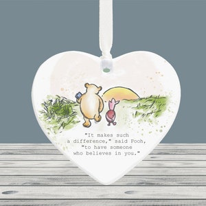 May include: Heart-shaped ornament with a watercolour illustration of Winnie the Pooh and Piglet. The ornament has a white background and a ribbon for hanging. Text reads: "It makes such a difference, to have someone who believes in you."