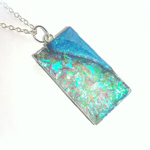 May include: A rectangular pendant necklace with a silver chain. The pendant features a shimmering, iridescent design with blue glitter on the top half. The bottom half of the pendant has a colorful, textured appearance.
