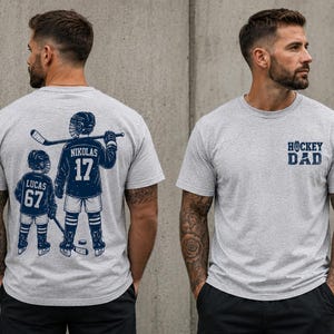 May include: Light gray t-shirt with a navy blue hockey-themed graphic. The front of the shirt has the text "HOCKEY DAD". The back of the shirt features a hockey player and a child with the names and numbers "NIKOLAS 17" and "LUCAS 67".