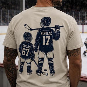 May include: A beige t-shirt with a navy blue hockey player graphic on the back, featuring the name "NIKOLAS" and the number "17", and a smaller player with the name "LUCAS" and the number "67". A black "HOCKEY DAD" tumbler is visible.