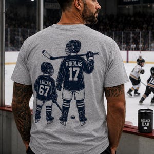 May include: A light grey t-shirt with a hockey-themed graphic on the back. The graphic shows two hockey players, one with the name "NIKOLAS" and the number "17", and the other with the name "LUCAS" and the number "67". A black container with the text "HOCKEY DAD" is also visible.