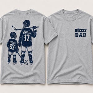 May include: Two gray t-shirts. One features a navy blue graphic of two hockey players with the names "Nikolas" and "Lucas" and the numbers 17 and 67. The other shirt has the text "HOCKEY DAD" in navy blue.