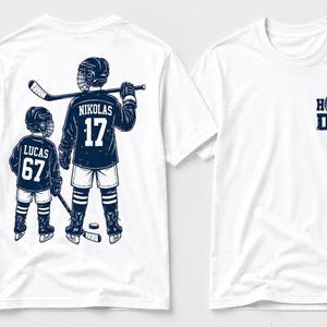 May include: White t-shirts with a hockey theme. One shirt features a graphic of two hockey players with the names "Nikolas" and "Lucas" and the numbers "17" and "67". The other shirt has the text "HOCKEY DAD".