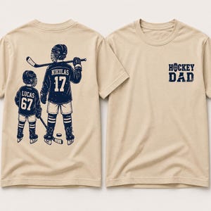 May include: Beige t-shirts with hockey-themed graphics. One shirt features a design of two hockey players with the text "NIKOLAS 17" and "LUCAS 67". The other shirt has the text "HOCKEY DAD" in navy blue.