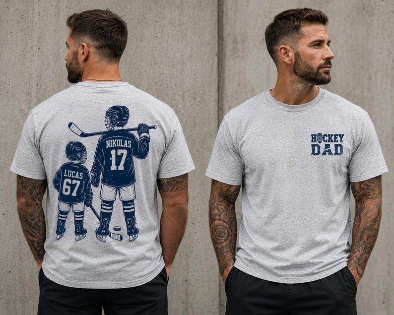 May include: Light gray t-shirt with a navy blue hockey-themed graphic. The front of the shirt has the text "HOCKEY DAD". The back of the shirt features a hockey player and a child with the names and numbers "NIKOLAS 17" and "LUCAS 67".