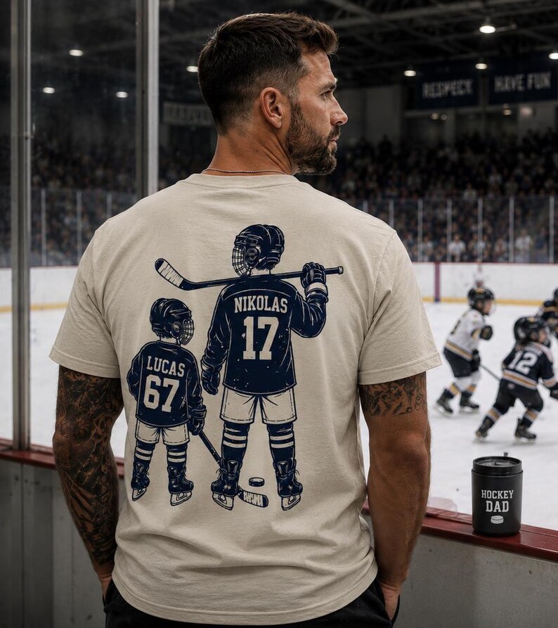 May include: A beige t-shirt with a navy blue hockey player graphic on the back, featuring the name "NIKOLAS" and the number "17", and a smaller player with the name "LUCAS" and the number "67". A black "HOCKEY DAD" tumbler is visible.