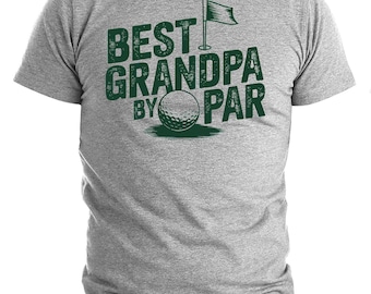 Puede incluir: Una camiseta gris con el texto verde "BEST GRANDPA BY PAR", una pelota de golf y un gráfico de bandera. La camiseta está hecha de un material suave y tiene mangas cortas.