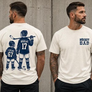 May include: Cream-colored t-shirt with a navy blue hockey-themed graphic. The front of the shirt says "HOCKEY DAD". The back of the shirt features a hockey player and a child with the names "NIKOLAS 17" and "LUCAS 67".
