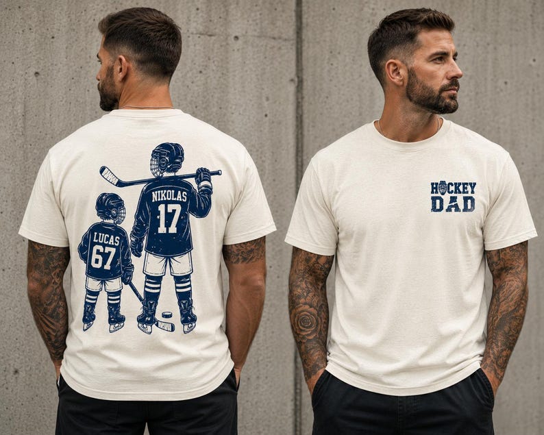 May include: Cream-colored t-shirt with a navy blue hockey-themed graphic. The front of the shirt says "HOCKEY DAD". The back of the shirt features a hockey player and a child with the names "NIKOLAS 17" and "LUCAS 67".