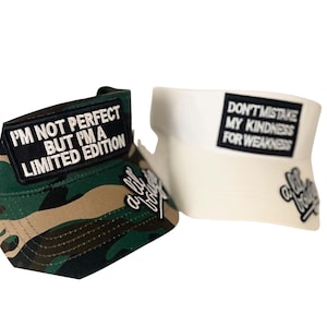 May include: Two visors with text. One is camouflage with the text "I'M NOT PERFECT BUT I'M A LIMITED EDITION". The other is white with the text "DON'T MISTAKE MY KINDNESS FOR WEAKNESS". Both visors have the text "a lil bolle".