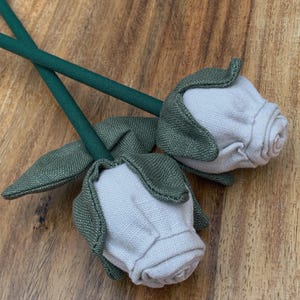 May include: Two fabric roses with light grey petals and green leaves and stems. The roses are made of a soft, textured material and are arranged on a wooden surface. The stems are a dark green colour.