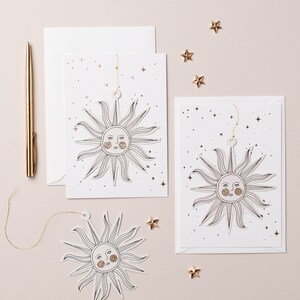 May include: Two white cards with gold glitter stars and a black line drawing of a sun with a face. The sun is cut out and attached to a string. The cards are on a light beige surface with gold glitter stars scattered around.