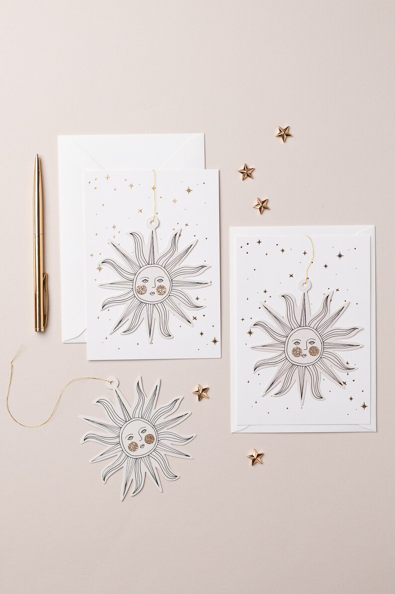 May include: Two white cards with gold glitter stars and a black line drawing of a sun with a face. The sun is cut out and attached to a string. The cards are on a light beige surface with gold glitter stars scattered around.
