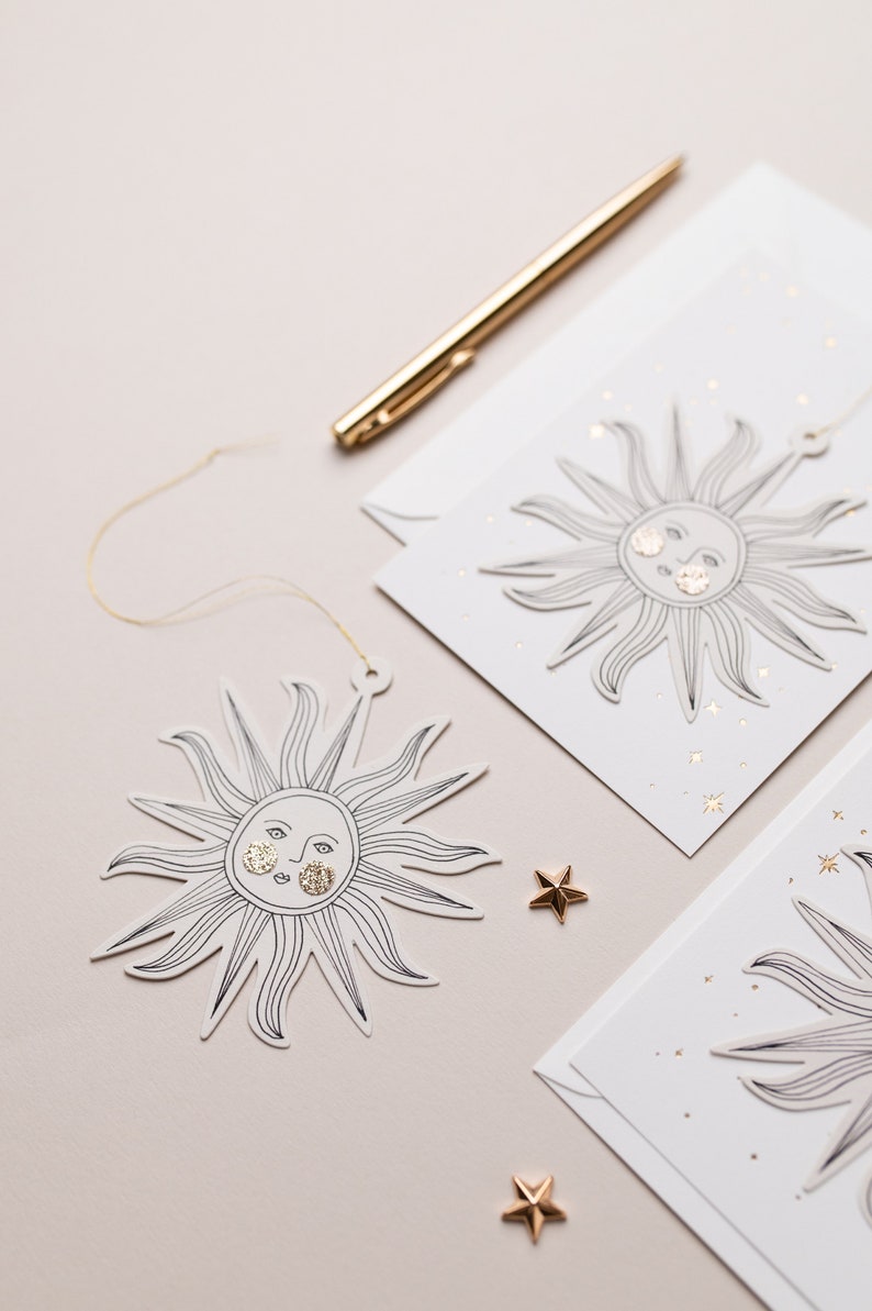 May include: Two white cards with gold glitter stars and a sun with a face drawn on them. One sun is hanging from a gold string. The cards are on a light pink surface.
