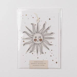 May include: A white card with a gold glitter sun cutout with a face. The card has a gold glitter background with stars. The card says "Handfinished Papercut Decoration with postcard & envelope".
