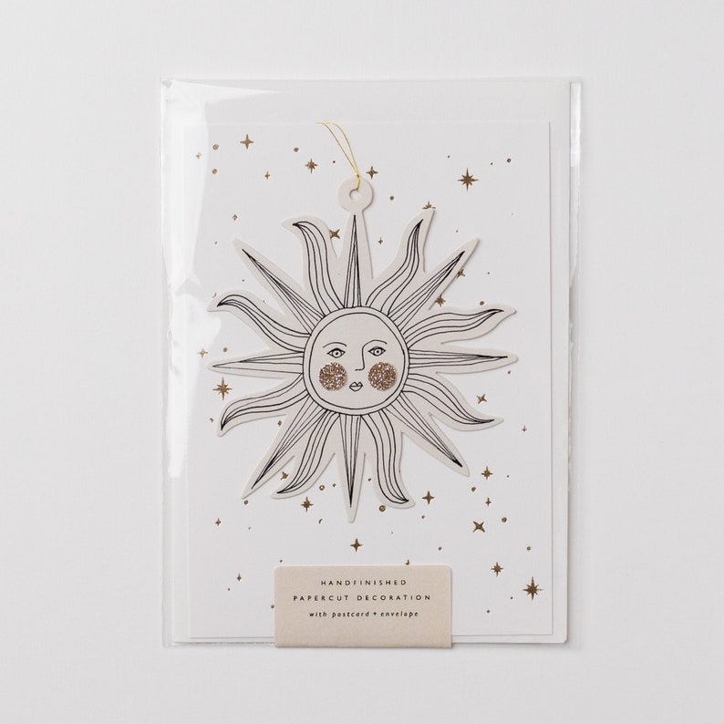 May include: A white card with a gold glitter sun cutout with a face. The card has a gold glitter background with stars. The card says "Handfinished Papercut Decoration with postcard & envelope".
