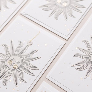 May include: Set of four white gift tags with gold foil accents. Each tag features a black line drawing of a sun with a face and a scattering of gold stars.