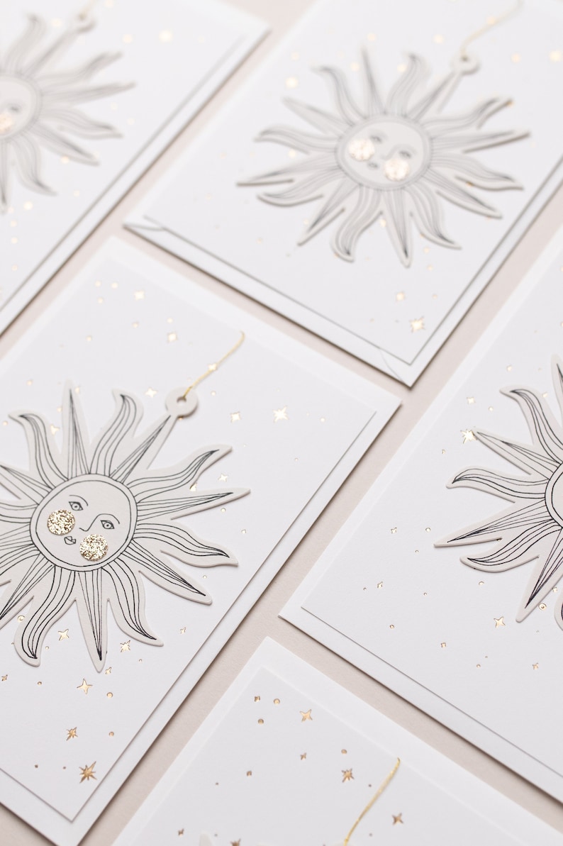 May include: Set of four white gift tags with gold foil accents. Each tag features a black line drawing of a sun with a face and a scattering of gold stars.