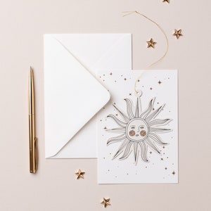 May include: A white card with a gold sun illustration and gold glitter stars. The card is tied with a gold string and is sitting on a light pink surface with a gold pen and gold stars.