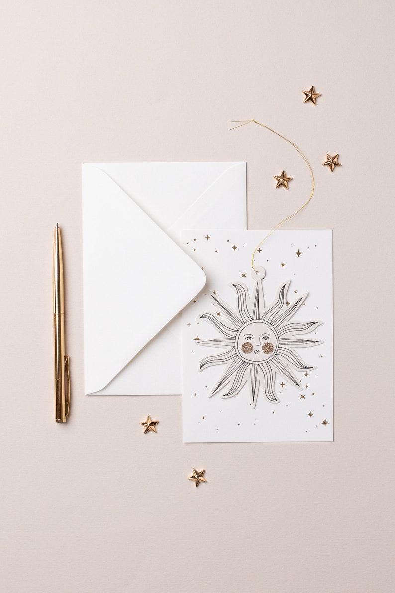 May include: A white card with a gold sun illustration and gold glitter stars. The card is tied with a gold string and is sitting on a light pink surface with a gold pen and gold stars.